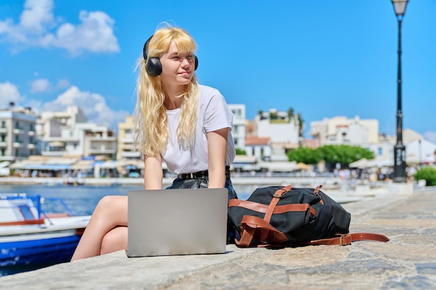 Adolescente femminile in cuffia con computer portatile seduto vicino al porto di mare con barche