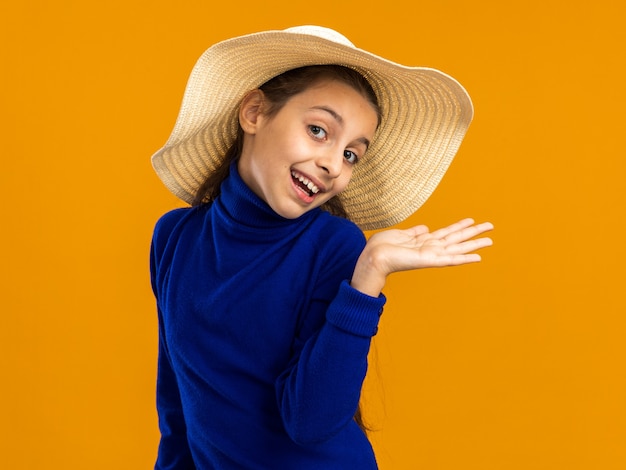 Adolescente gioiosa che indossa il cappello da spiaggia che mostra la mano vuota guardando la parte anteriore isolata sulla parete arancione