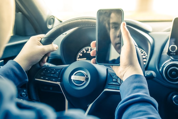 Adolescente guida un'auto e usa uno smartphone giovane che legge messaggi con un cellulare in mano mentre guida