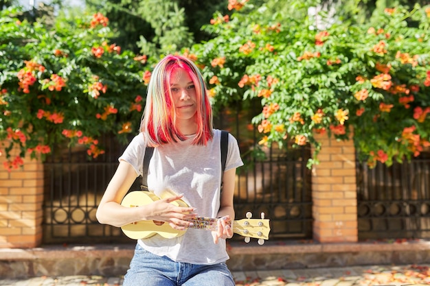 Adolescente hipster che suona l'ukulele seduto all'aperto, soleggiata giornata estiva in città. Ragazza di tendenza creativa con i capelli tinti con ukulele, musica, hobby e svago della giovinezza