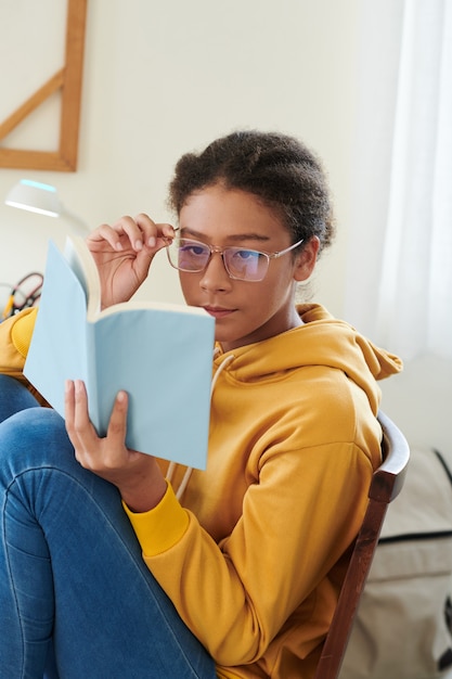 Adolescente intelligente di razza mista focalizzato seduto con i piedi sulla sedia e regolando gli occhiali mentre legge il libro a casa