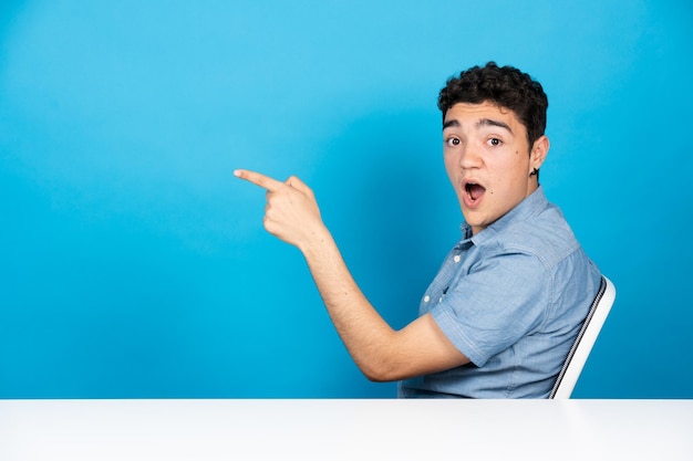 Adolescente ispanico seduto che punta allo spazio della copia isolato su sfondo blu