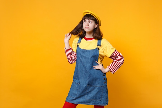 Adolescente pensieroso della ragazza in prendisole del denim del berretto francese che sta con il braccio akimbo che tiene i capelli isolati sul fondo giallo della parete in studio. Persone sincere emozioni, concetto di stile di vita. Mock up spazio di copia.