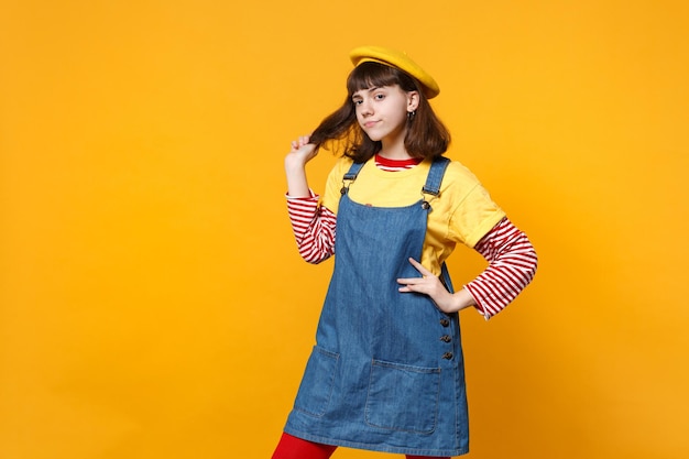 Adolescente perplesso della ragazza in prendisole del denim del berretto francese che sta con il braccio akimbo che tiene i capelli isolati sul fondo giallo della parete in studio. Persone sincere emozioni, concetto di stile di vita. Mock up spazio di copia.