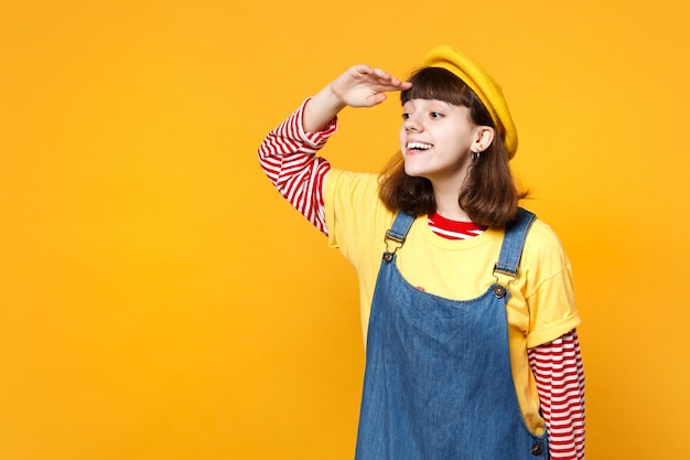 Adolescente ragazza allegra in berretto francese, prendisole in denim tenendo la mano sulla fronte guardando lontano distanza isolata su sfondo giallo. Persone sincere emozioni, concetto di stile di vita. Mock up spazio di copia.