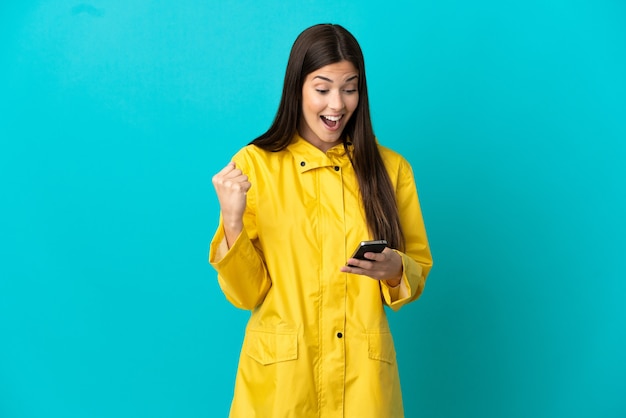 Adolescente ragazza brasiliana che indossa un cappotto antipioggia su sfondo blu isolato sorpreso e l'invio di un messaggio