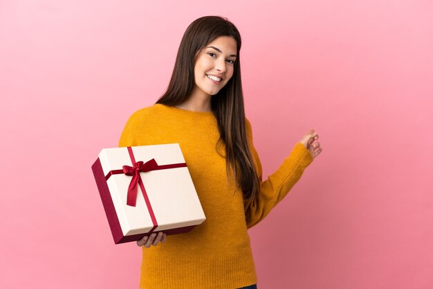 Adolescente ragazza brasiliana che tiene un regalo su sfondo rosa isolato che punta indietro