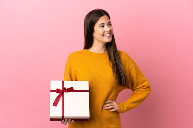 Adolescente ragazza brasiliana che tiene un regalo su sfondo rosa isolato in posa con le braccia all'anca e sorridente