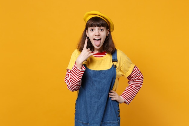 Adolescente ragazza divertente in berretto francese che mostra la lingua, il gesto delle corna, raffigurante il segno rock di metalli pesanti, rock-n-roll isolato su sfondo giallo. Concetto di stile di vita delle emozioni delle persone. Mock up spazio di copia.