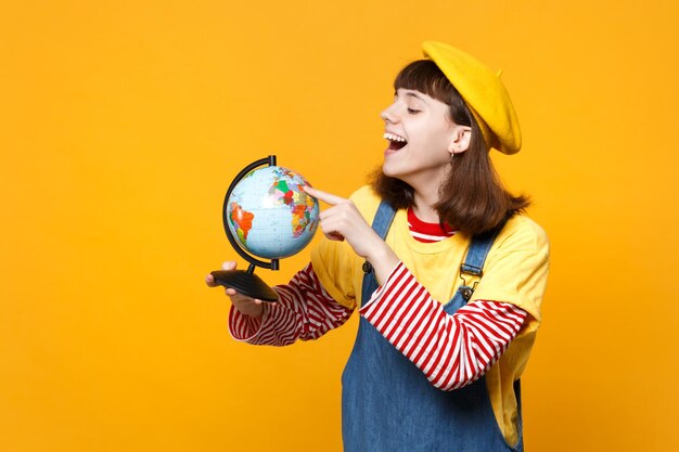 Adolescente ragazza ridente in berretto francese e prendisole in denim che punta il dito indice sul globo terrestre isolato su sfondo giallo muro. Persone sincere emozioni, concetto di stile di vita. Mock up spazio di copia.