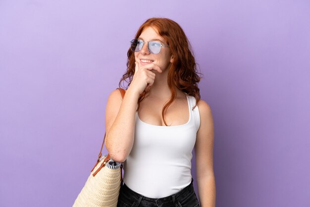 Adolescente ragazza rossa che tiene una borsa da spiaggia isolata su sfondo viola guardando al lato