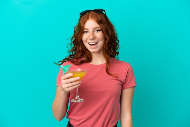 Adolescente ragazza rossa con cocktail isolato su sfondo blu con sorpresa e espressione facciale scioccata