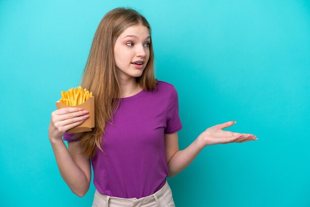 Adolescente ragazza russa che cattura patatine fritte isolate su sfondo blu con espressione facciale a sorpresa