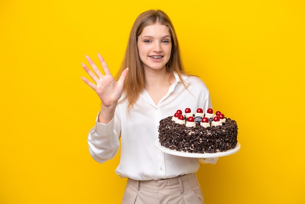 Adolescente ragazza russa che tiene la torta di compleanno isolata su sfondo giallo contando cinque con le dita