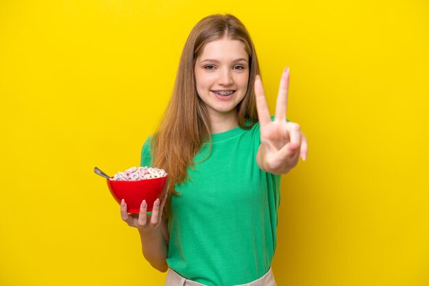 Adolescente ragazza russa con ciotola di cereali isolata su sfondo giallo sorridente e mostrando il segno di vittoria