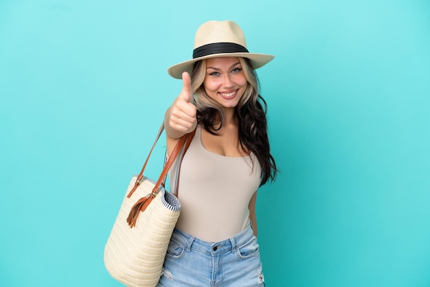 Adolescente ragazza russa con pamel e borsa da spiaggia isolata su sfondo blu con i pollici in su perché è successo qualcosa di buono