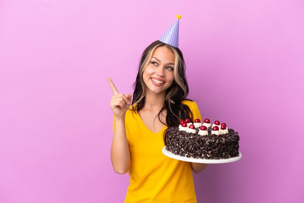 Adolescente ragazza russa con torta di compleanno isolata su sfondo viola con l'intenzione di realizzare la soluzione sollevando un dito