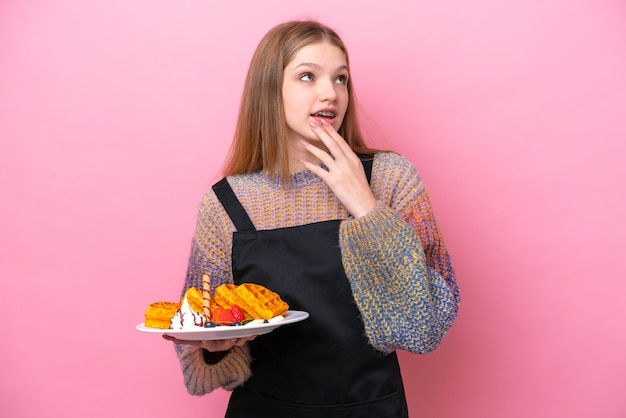 Adolescente ragazza russa in possesso di un waffle isolato su sfondo rosa guardando in alto mentre sorride