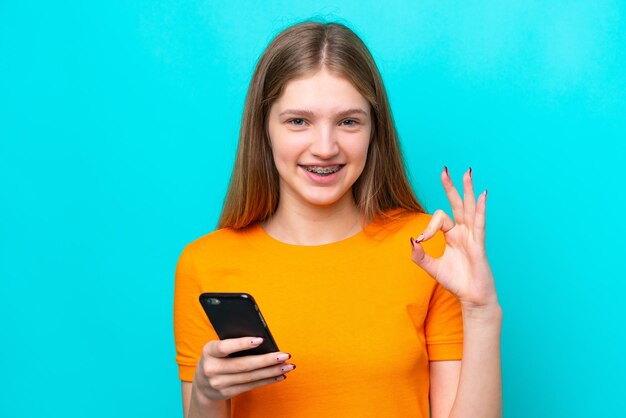 Adolescente ragazza russa isolata su sfondo blu utilizzando il telefono cellulare e facendo segno OK