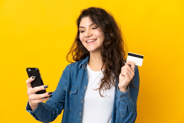 Adolescente ragazza russa isolata su sfondo giallo che acquista con il cellulare con una carta di credito