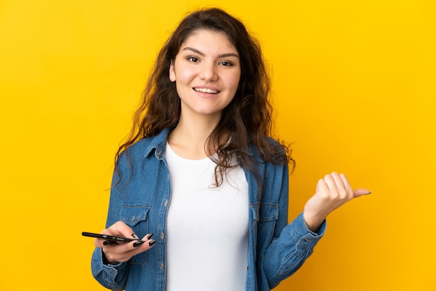 Adolescente ragazza russa isolata su sfondo giallo utilizzando il telefono cellulare e che punta al laterale