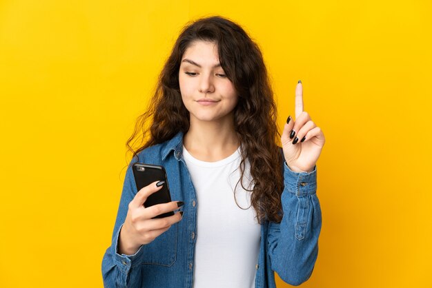 Adolescente ragazza russa isolata su sfondo giallo utilizzando il telefono cellulare e sollevando il dito