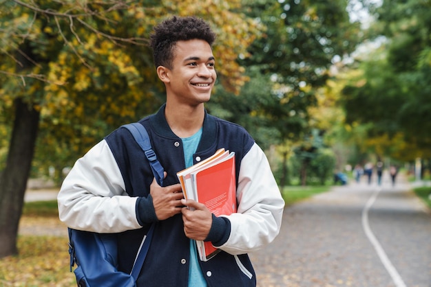 Adolescente ragazzo allegro che cammina al parco, portando zaino e libri di testo