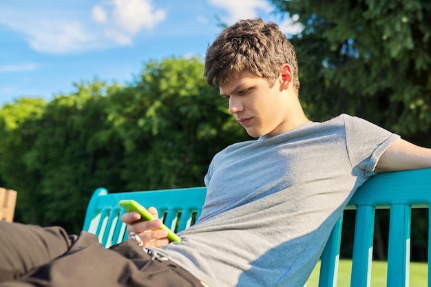 Adolescente ragazzo serio che utilizza smartphone riposando seduto su una panchina nel parco