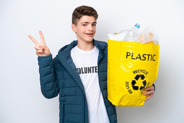 Adolescente russo in possesso di un sacchetto pieno di bottiglie di plastica da riciclare su sfondo bianco sorridente e mostrando il segno della vittoria