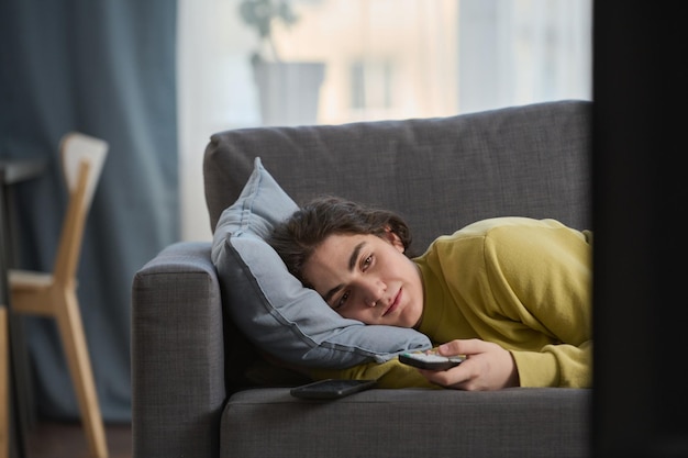 Adolescente sdraiato sul divano e cambiando canale con telecomando mentre si guarda la tv in camera
