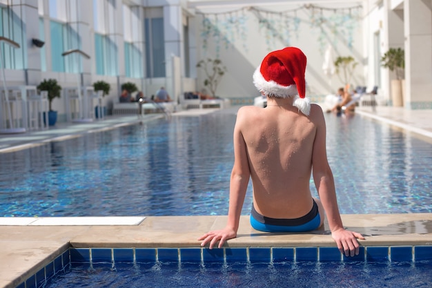 Adolescente senza volto in cappello rosso di Babbo Natale seduto sul bordo della piscina in una giornata di sole