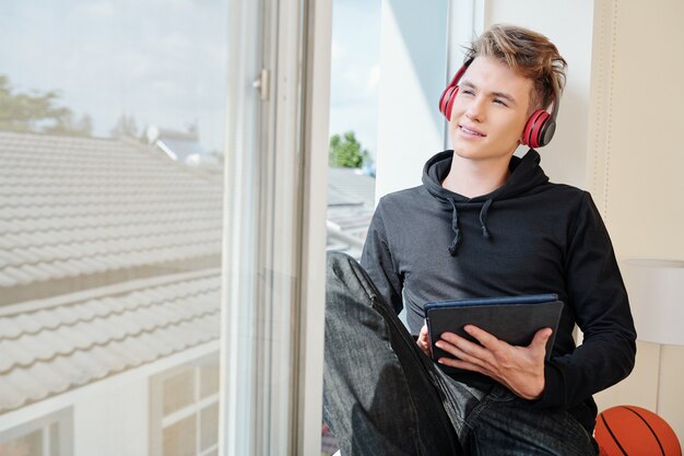 Adolescente sorridente con cuffie e tavoletta digitale seduto sul davanzale della finestra e guardando il cielo quando si resta a casa durante il blocco