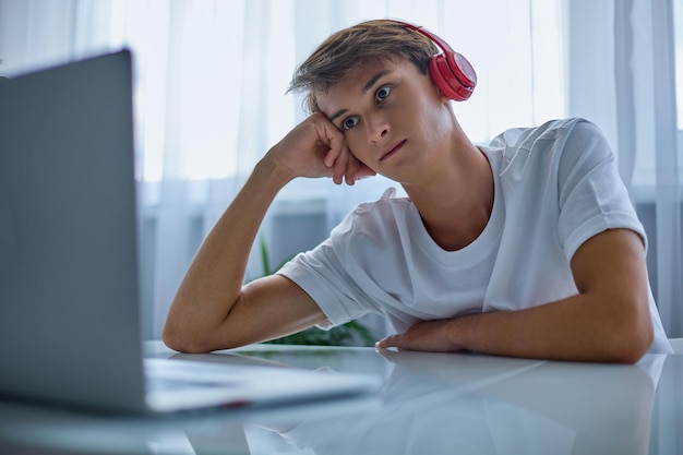 Adolescente stanco in cuffie rosse sta guardando un computer portatile