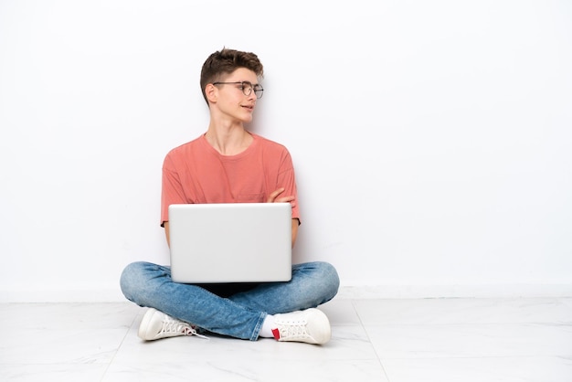 Adolescente uomo russo azienda pc seduto sul pavimento isolato su sfondo bianco guardando lato