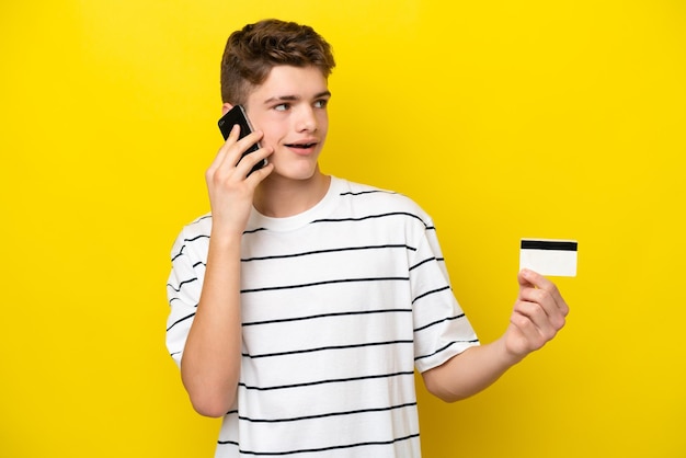 Adolescente uomo russo isolato su sfondo giallo mantenendo una conversazione con il telefono cellulare e tenendo una carta di credito