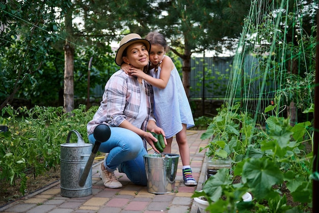 Adorabile bambina abbraccia sua madre mentre raccoglie cetrioli maturi nel giardino ecologico della casa di campagna Agroalimentare familiare