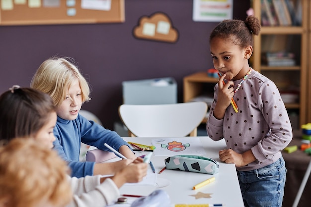 Adorabile bambina afroamericana con i pastelli che guardano un gruppo di compagni di classe seduti davanti o