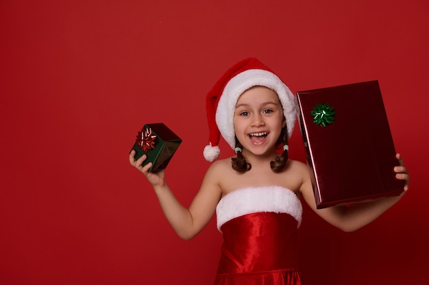 Adorabile bambina allegra in abiti da carnevale di Babbo Natale, tiene in mano una scatola regalo di Natale in carta da imballaggio verde e rossa e gioisce sorridendo alla telecamera in posa su sfondo colorato con spazio copia per annuncio
