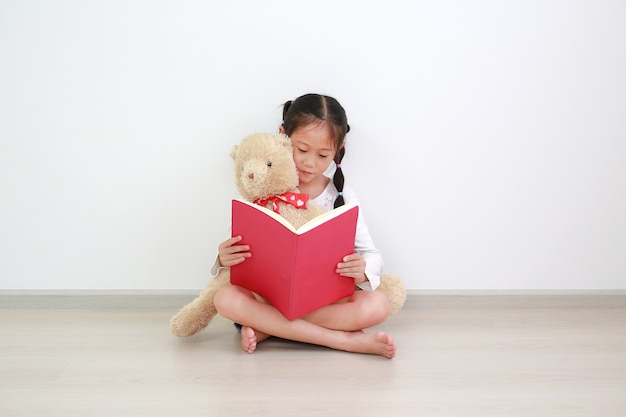 Adorabile bambina asiatica bambina che legge un libro con abbracciare una bambola di orsacchiotto seduto contro il muro bianco nella stanza.