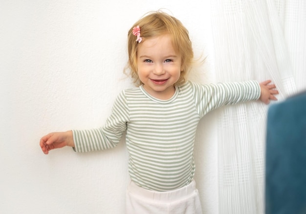 Adorabile bambina bionda caucasica di 12 anni che gioca con tende bianche divertendosi a casa in interni luminosi guardando la telecamera