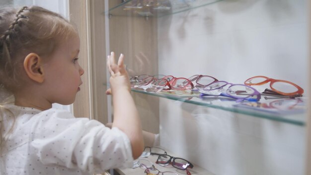 Adorabile bambina bionda nella clinica di oftalmologia gioca nel corridoio vicino al negozio di occhiali, primo piano