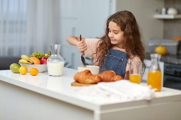 Adorabile bambina carina con i capelli ricci che mangia una sana colazione a base di cereali in cucina.