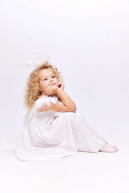 Adorabile bambina carina in abito bianco e aureola di angeli seduta con il viso sognante isolato