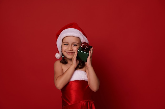Adorabile bambina carina, splendida bambina in abiti da carnevale di Babbo Natale abbraccia delicatamente la sua confezione regalo di Natale in carta da regalo verde glitterata e fiocco rosso, sorride graziosamente guardando la fotocamera. Copia spazio per l'annuncio