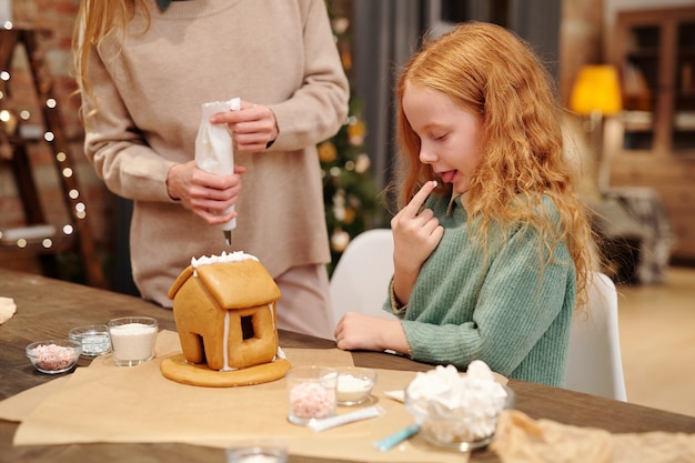 Adorabile bambina che assaggia la panna montata dal tetto della casa di marzapane fatta in casa mentre sua madre la decora prima della cena di Natale