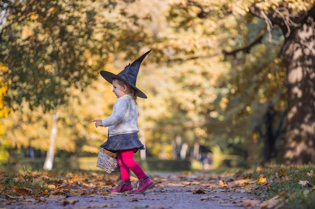 adorabile bambina che cammina nel parco autunnale. ragazza in costume da strega e cappello nero cosplay costume di halloween. festa di halloween