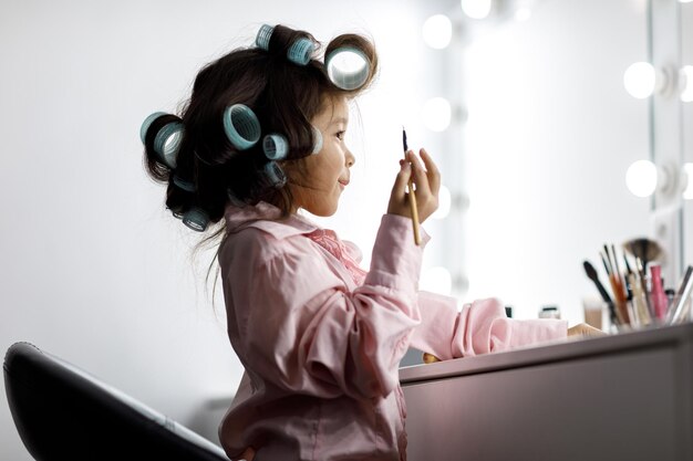 Adorabile bambina che gioca con il cosmetico di sua madre davanti allo specchio a casa