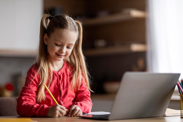 Adorabile bambina che scrive appunti mentre studia con il computer portatile a casa