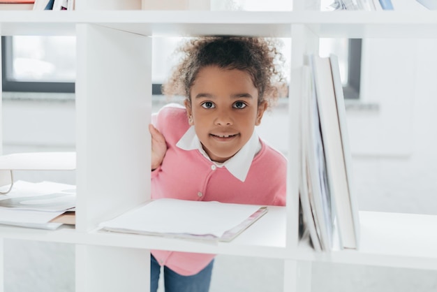 Adorabile bambina che sorride alla macchina fotografica attraverso gli scaffali in ufficio