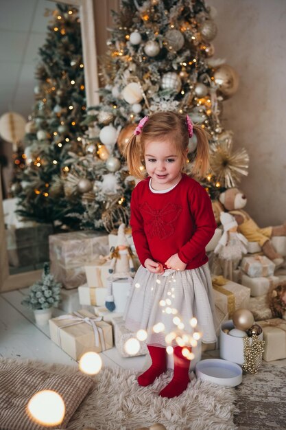 Adorabile bambina che tiene le luci della ghirlanda sullo sfondo dell'albero di Natale